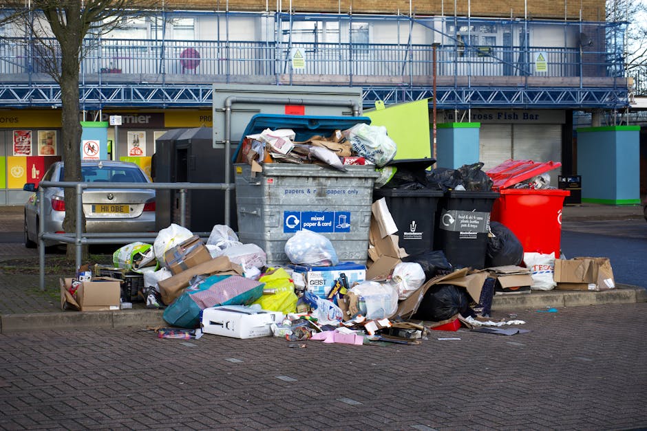 A large, unmanaged pile of mixed waste and rubbish, consisting of various materials such as cardboard boxes, plastic bags, paper, and household items, is overflowing from multiple black, red, and grey