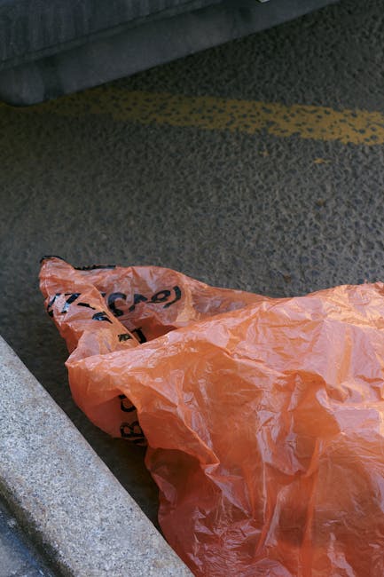 A narrow urban alleyway cluttered with various waste materials. In the center, a large, partially transparent black and grey bin bag filled with rubbish is positioned on uneven asphalt ground, showing