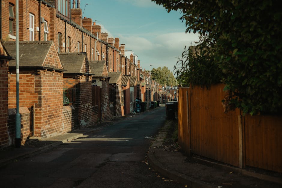 A residential street featuring detached houses with brick walls, white window frames, and grey tiled roofs. The houses are fronted by a narrow pavement and a road with parked cars, including a dark gr